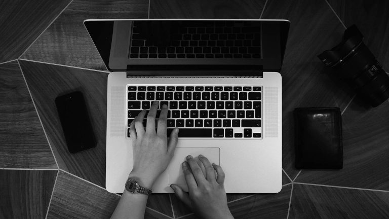 Black and white top-down view of a person's hands typing on a laptop, with a camera, smartphone, and wallet on the desk.