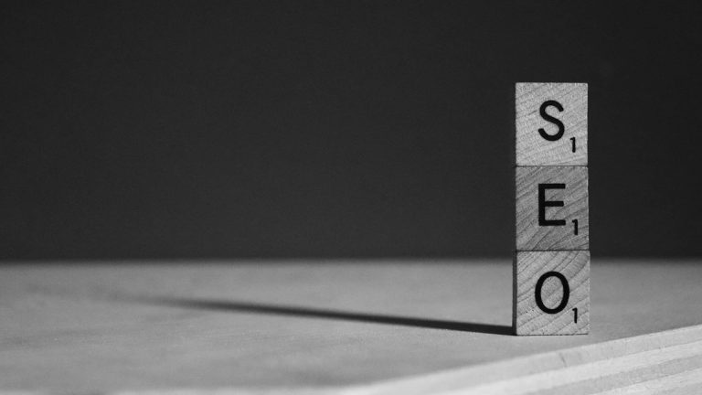 Black and white photo of three wooden Scrabble tiles stacked vertically to spell out the acronym "SEO".