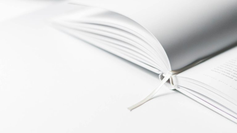 A close-up of an open book with a ribbon bookmark, resting on a white surface.