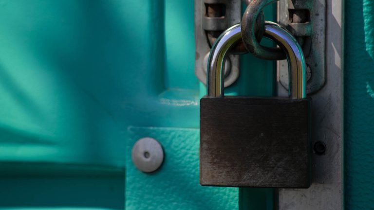Close-up of a silver and black padlock securing a chain on a teal-colored door.
