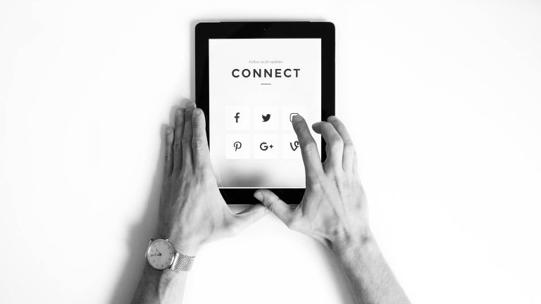 Black and white top-down view of hands holding a tablet, with a finger pointing to the Instagram icon on a "Connect" page.