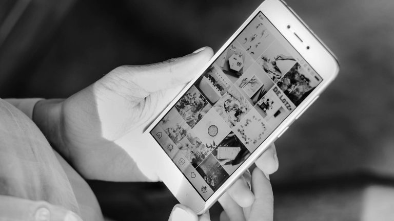 Black and white photo of a person's hands holding a smartphone and scrolling through an Instagram profile feed.