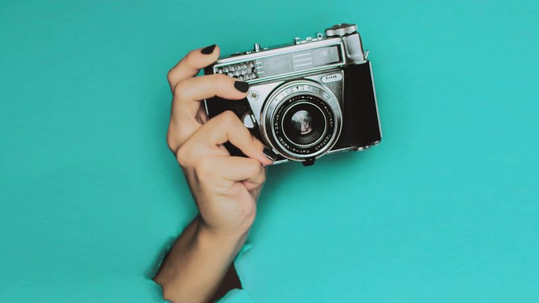 A hand with black nail polish holding a vintage film camera, emerging from a hole in a teal background.