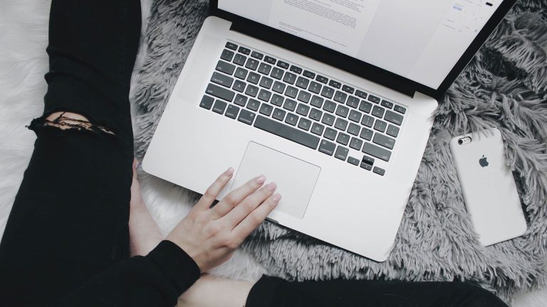 Top-down view of a person sitting on a shaggy grey rug using a laptop, with a smartphone nearby.