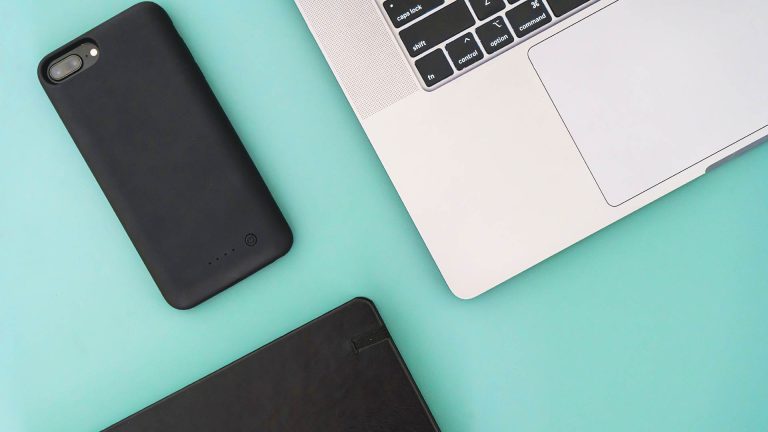 Top-down flat lay of a silver laptop, a black smartphone, and a notebook on a mint green desk.