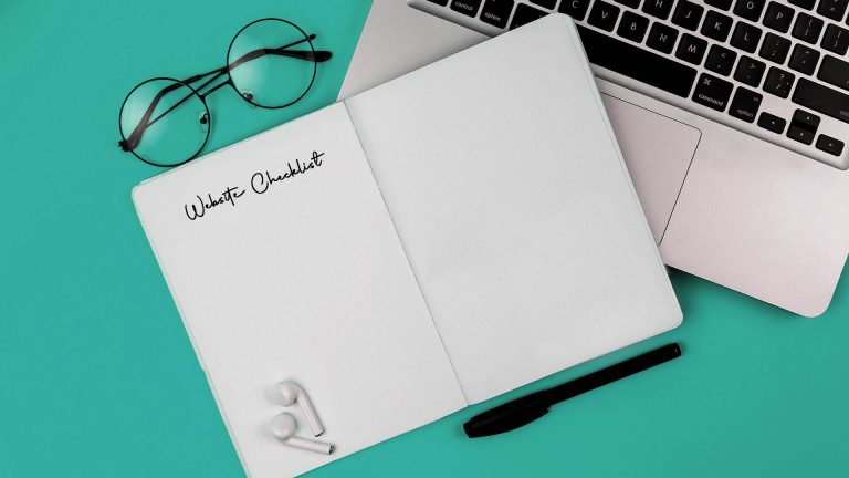 A flat lay of an open notebook with "Website Checklist" written on it, next to a laptop, glasses, and earbuds.